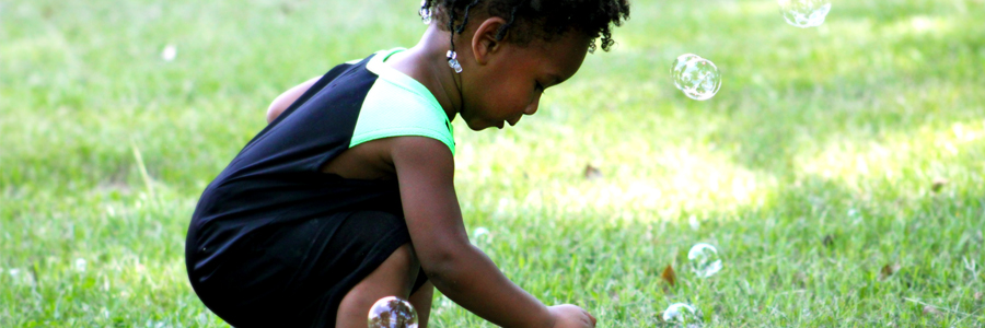 Fotografia de uma garotinha vestindo um macacão preto com verde, agachada sobre a grama brincando bolhas de sabão.